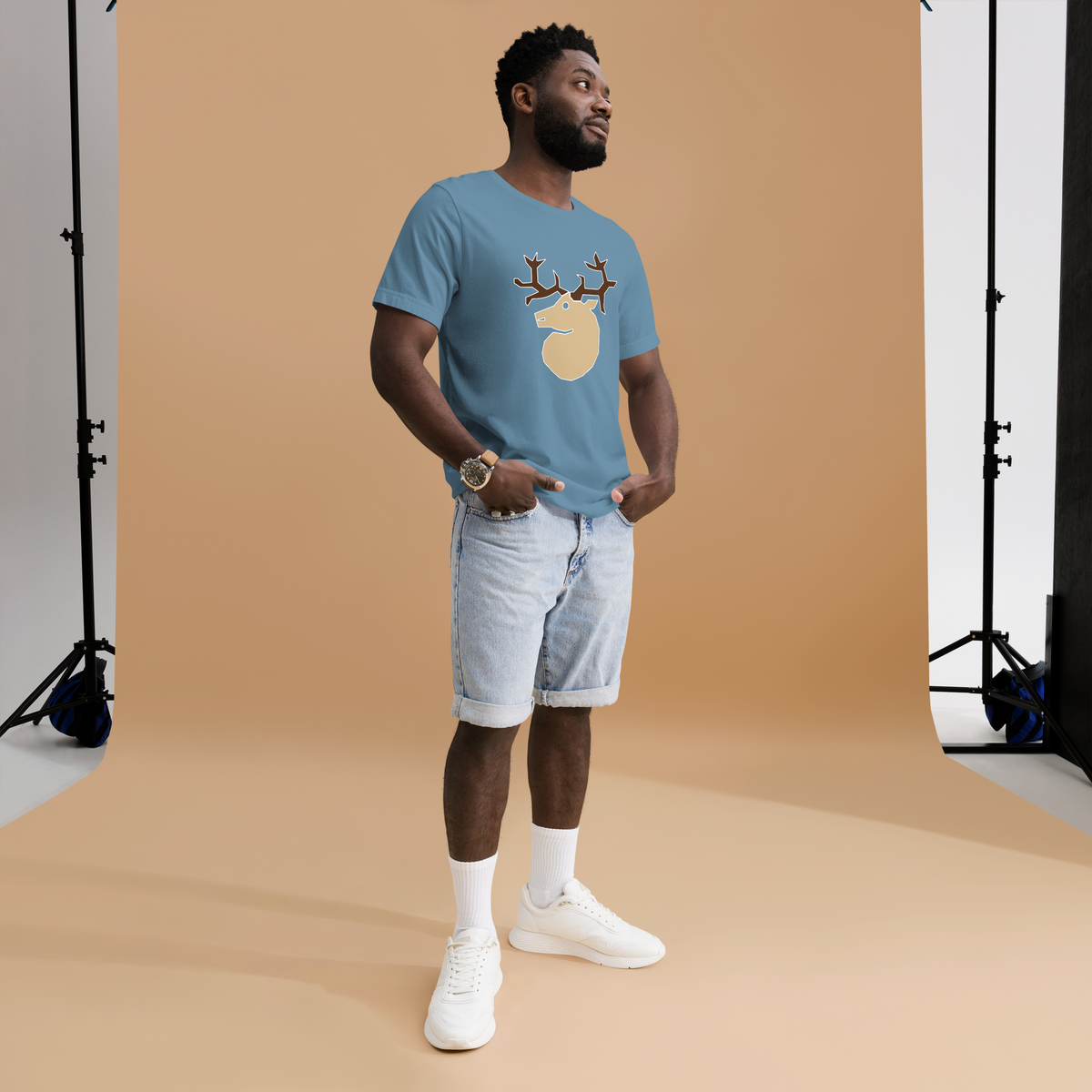 Fashionable male model against brightly lit orange background wearing a steel blue 100% airlume cotton t-shirt featuring classic fan-made woodland outlaws deer / stag crest design. Item is a BRKDrip AFOL essentials shirt with a vintage castle building blocks aesthetic. 