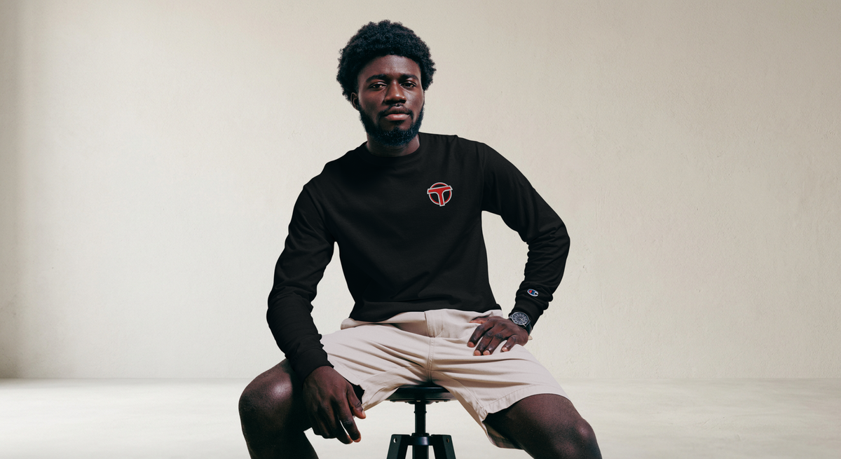 Fashionable male model sitting on a stool wearing a black lightweight, 100% cotton luxury Champion© shirt featuring airport shuttle emblem embroidered in the top right corner. Item is a BRKDrip Builder's Collection luxury longsleeve shirt with a classic town building block aesthetic.