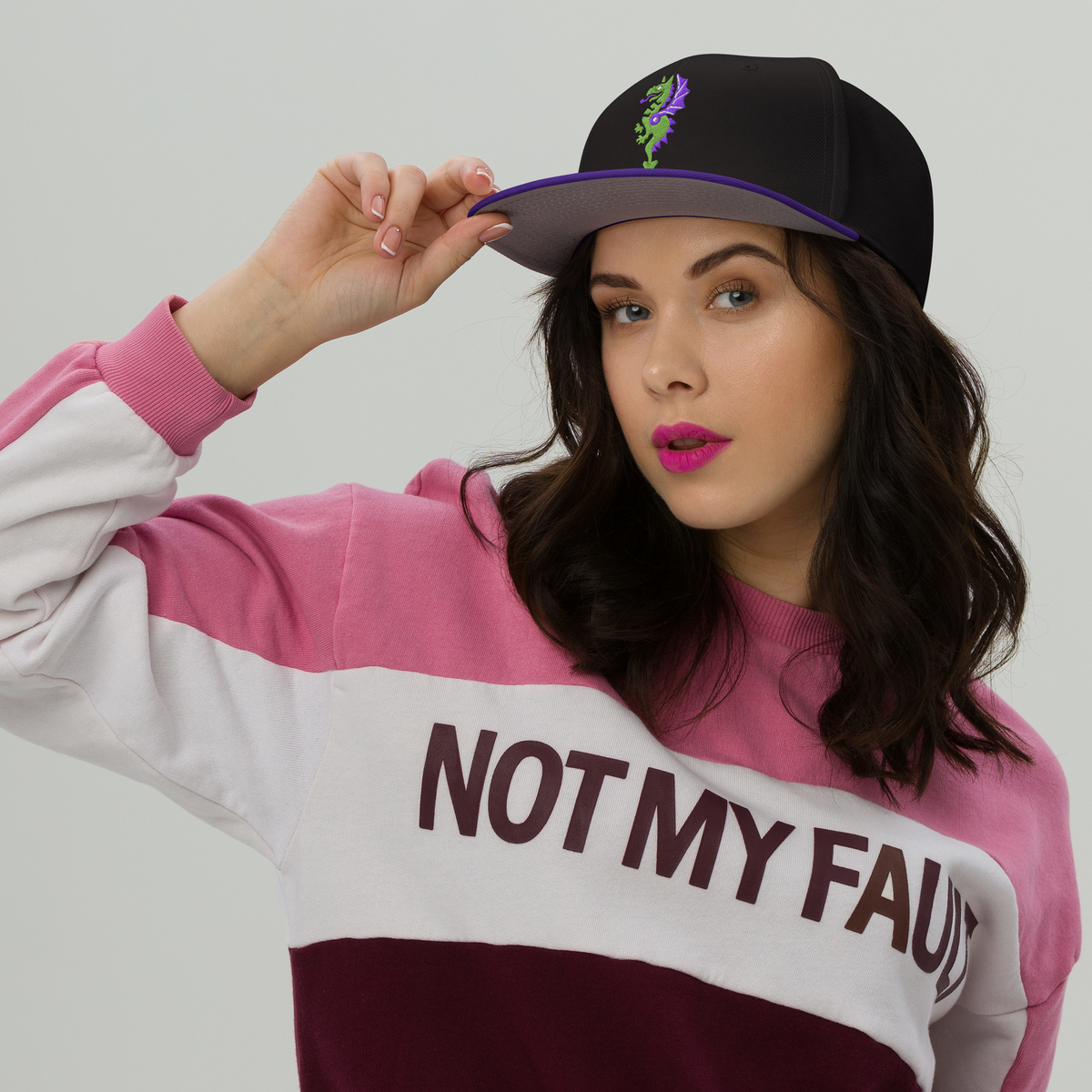 Fashionable female model wearing a black flat bill hat with a bright purple bill and strap featuring embroidered dragon master remix design with matching bright purple wings. Item is a BRKDrip AFOL Headgear classic snapback with a vintage castle building blocks aesthetic.